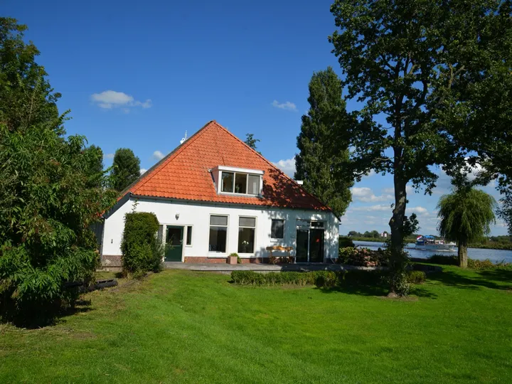 Vrijzicht vanaf de zuidkant: de witte woonboerderij met rode pannen, vrijstaand in het gras, met het kanaal erachter